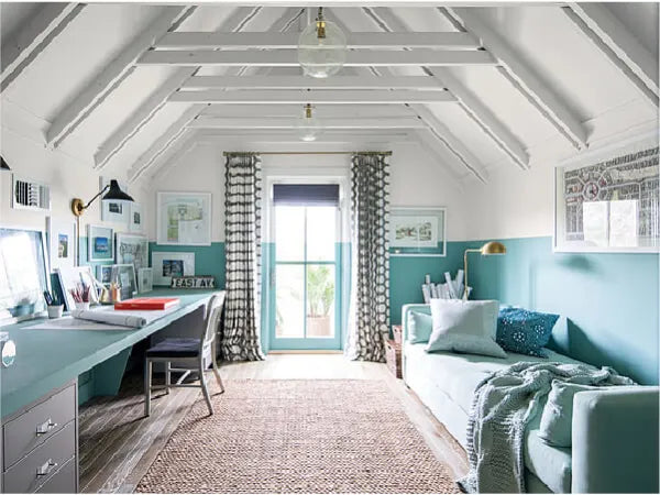 Bright attic room with sloped white ceiling beams, teal half walls, a long desk with office supplies, and a cozy daybed with cushions and a blanket