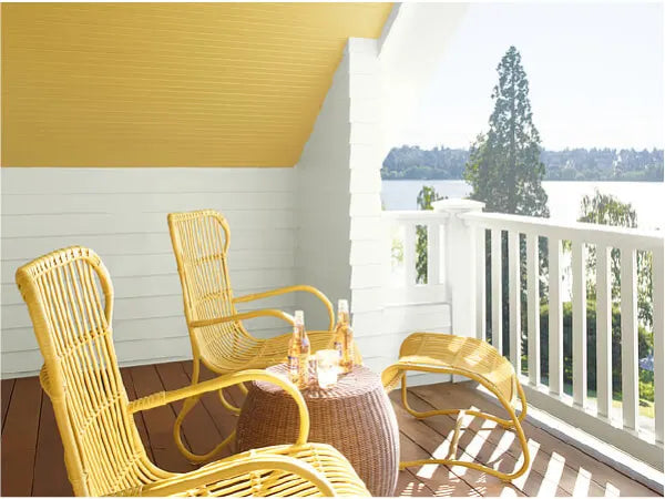 Sunny porch corner with yellow wicker chairs and matching ottoman surrounding a round woven table, overlooking a lake and trees beyond a white railing