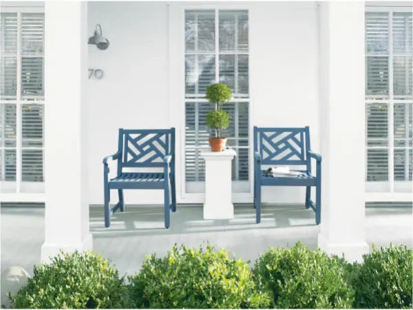 Front porch with two blue wooden chairs flanking a white pedestal holding a potted topiary, set against white walls and windows with blinds.