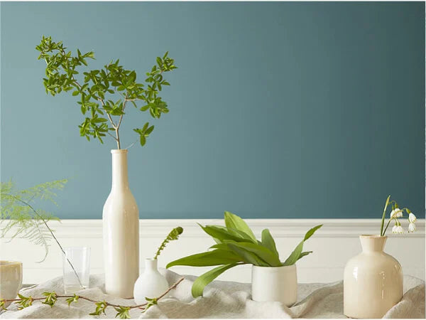 Arrangement of white ceramic vases and small plants on a linen-covered surface against a muted blue-green wall with white wainscoting.