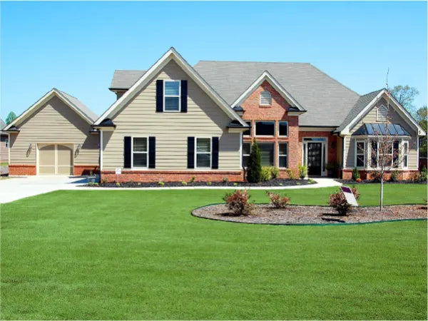 Large suburban house with beige siding and brick accents, surrounded by a neatly manicured green lawn and a clear blue sky.