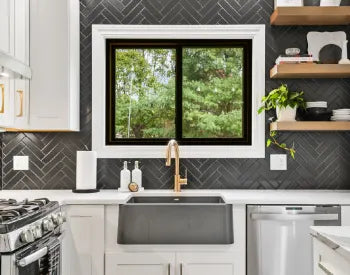 Modern kitchen sink area with a large black basin, brass faucet, white countertops, dark herringbone tile backsplash, and wooden floating shelves displaying decor.