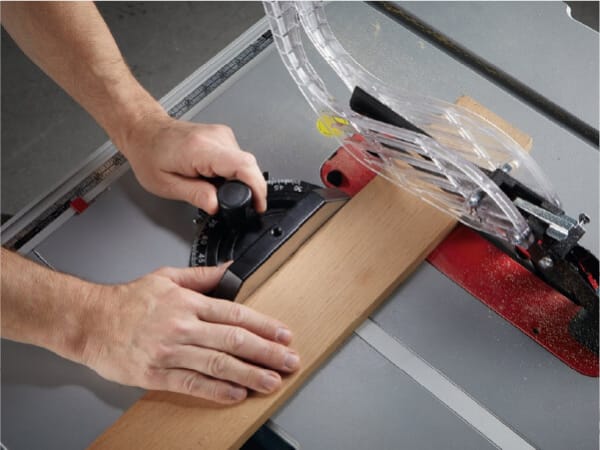 Person using a table saw to carefully cut a piece of wood, adjusting the fence with one hand and guiding the wood with the other, in a workshop setting.