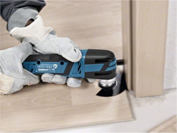 Person wearing gloves operates a blue and black cordless oscillating multi-tool to trim a wooden floor edge near a doorway during home improvement.