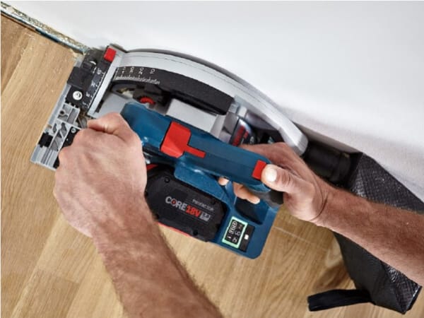 Person using a blue cordless circular saw to cut wooden flooring near a white wall, focusing on precise home improvement work.