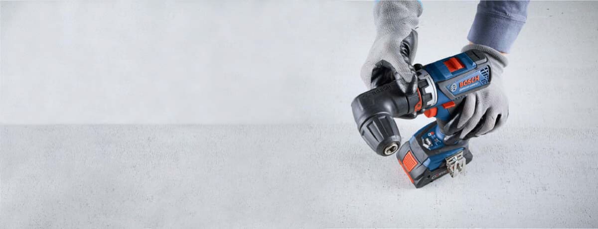 Person wearing gloves holding a compact blue cordless power drill with an angled attachment, positioned above a smooth light gray surface.