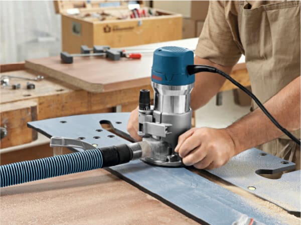 Person using a blue handheld router with a dust extraction hose attached to shape or trim a wooden surface in a workshop environment.