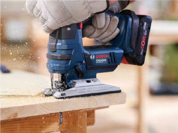 Close-up of a person wearing gloves using a blue cordless jigsaw to cut a piece of wood with sawdust flying, shown in a workshop setting.