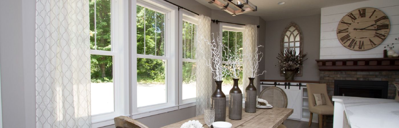 Cozy dining area with large white-framed windows letting in natural light, neutral curtains, wooden table with decorative vases, and a brick fireplace with a large clock above.