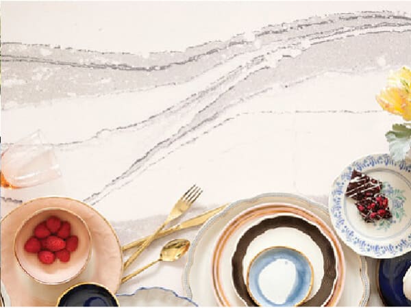 Light-colored stone surface with soft gray veining, featuring an arrangement of assorted decorative plates, gold flatware, and a small floral accent on the side.