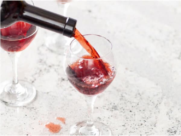 Red wine being poured from a bottle into a clear wine glass on a light-colored speckled countertop with a small spilled drop nearby.