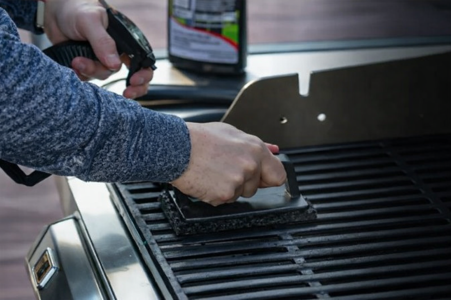 Close-up of hands cleaning a metal grill grate using a wire brush and spray bottle, with a wooden deck surface in the background.