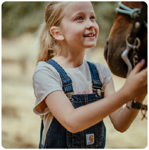 Young girl in denim overalls gently holding and looking up at a horse with a soft, natural outdoor background.