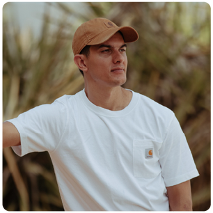 Man wearing a light brown cap and white short-sleeve shirt with a small logo on the pocket, standing outdoors with blurred greenery in the background.