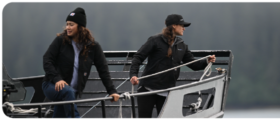 Two women dressed in black outdoor jackets and hats standing on a boat with a forested background, one holding a rope at the bow.