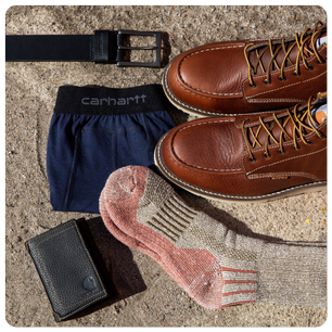 Pair of brown leather lace-up boots placed next to a black belt, navy blue underwear, a pair of cushioned socks, and a black leather wallet laid out on a textured surface.
