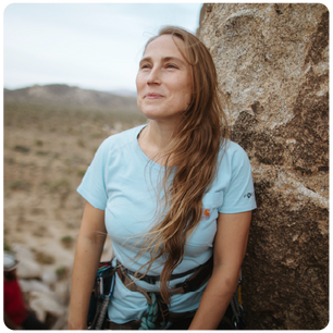 Woman with long light brown hair wearing a light blue t-shirt and climbing harness standing next to a rock in a desert landscape, looking slightly upward with a content expression.