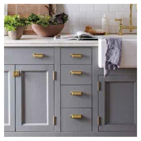 Kitchen cabinetry painted in muted gray with brass handles and hinges, topped with a white countertop, a farmhouse sink, and decorative plants nearby.