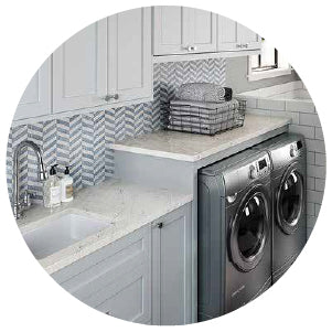 Bright laundry room featuring gray front-loading washer and dryer beneath a white quartz countertop with a modern herringbone tile backsplash and light gray cabinetry.