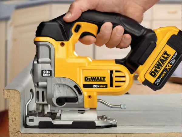Person's hand holding a yellow and black cordless jigsaw cutting through a piece of wood in a workshop setting.