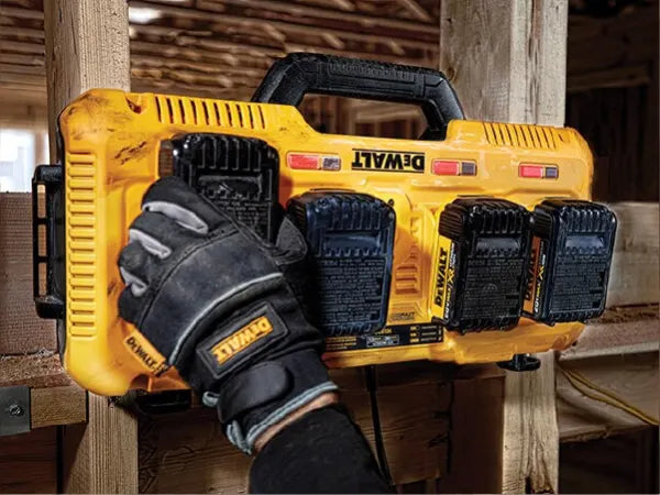 Gloved hand placing a black battery into a yellow tool charging station mounted on a wooden frame in a workshop environment.