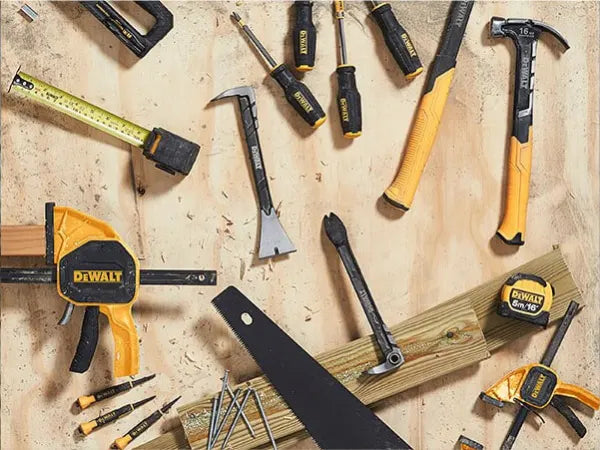 Collection of hand tools including hammers, screwdrivers, measuring tape, clamps, nails, and a saw arranged on a wooden surface.
