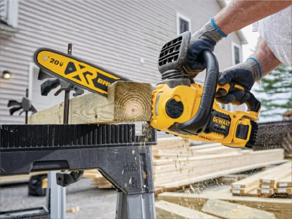 Person wearing gloves operating a yellow cordless chainsaw to cut a wooden beam outdoors near a house under construction.
