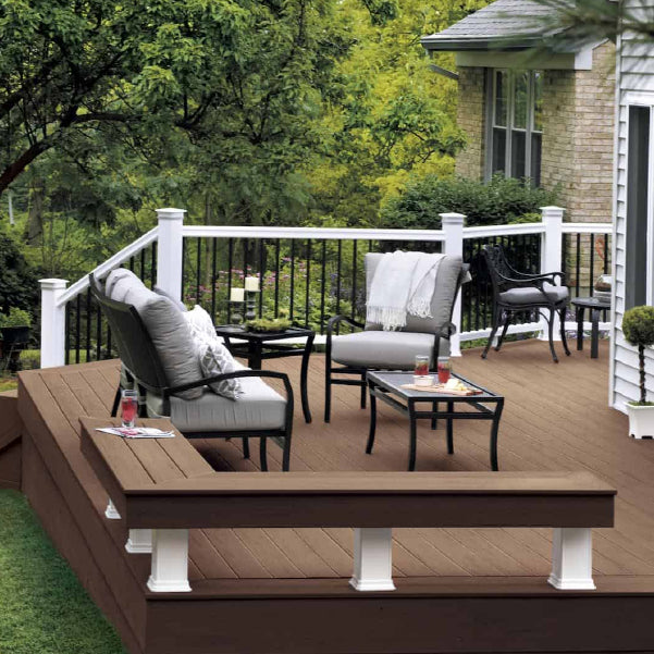 Outdoor wooden deck attached to a house showing cushioned chairs, a small table with drinks, and a railing overlooking green trees and bushes.