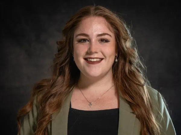 Portrait of a woman with long wavy hair wearing a light olive blazer and black top, smiling against a dark blurred background.