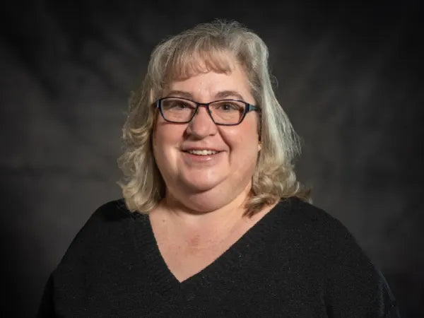 Middle-aged woman with light curly hair and glasses wearing a black top, smiling against a dark blurred background.