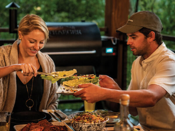 Two people sharing a meal outdoors on a wooden porch, passing a plate of grilled vegetables with a smoker grill visible in the background.