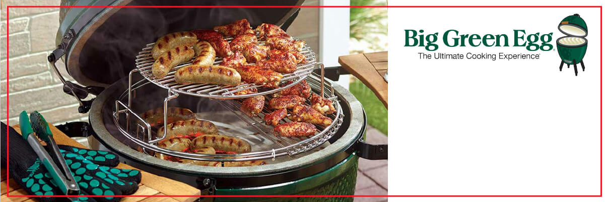 Green ceramic grill with the lid open showing chicken and sausages cooking on two-tiered racks, set on a patio table next to outdoor utensils.
