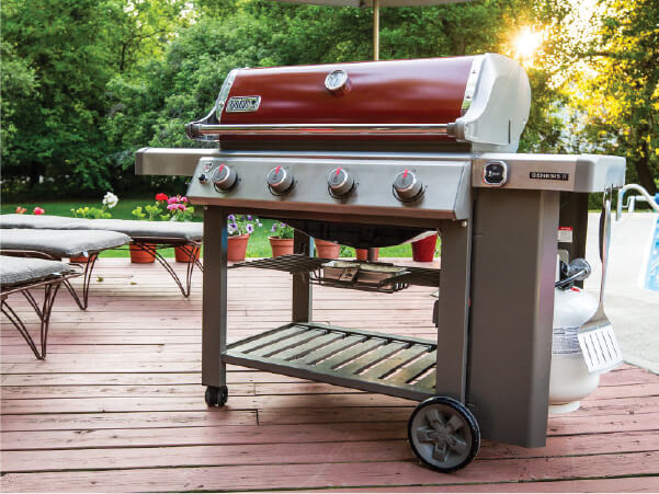 Four-burner gas grill with a red lid and stainless steel body standing on a wooden deck surrounded by patio furniture and greenery in soft sunlight.