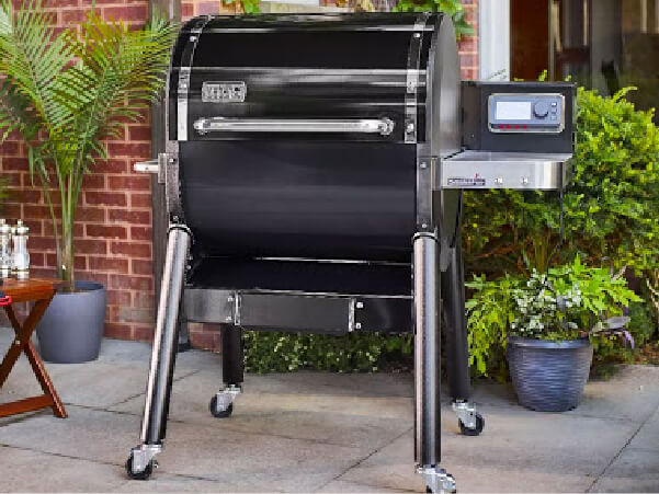 Black outdoor pellet grill with digital control panel on the side, standing on a patio with potted plants and a brick wall backdrop.
