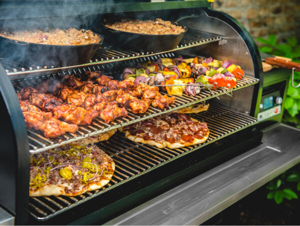 Open grill filled with seasoned chicken wings, vegetable skewers, and pizzas cooking on multiple racks in an outdoor setting