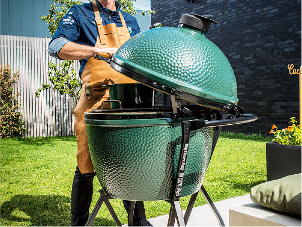 Person wearing a leather apron lifting the lid of a large green ceramic charcoal grill on a sunny backyard patio.