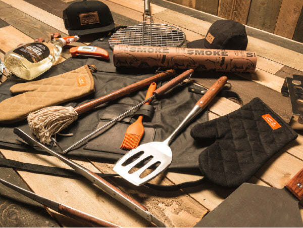 Assortment of grilling tools and accessories including a spatula, tongs, basting brush, gloves, apron, charcoal, hats, and a bottle on a wooden table.