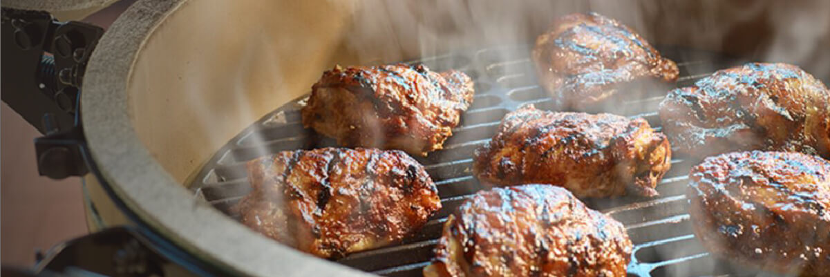 Close-up of several pieces of seasoned chicken cooking on a ceramic grill with visible smoke rising in a warm indoor setting.