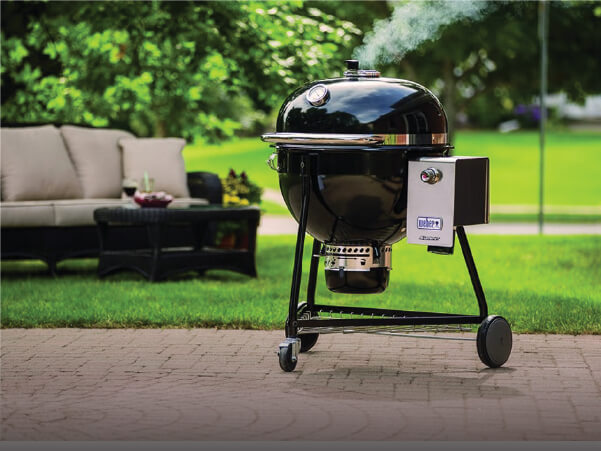 Black spherical charcoal grill with smoking vent open and attached smoker box, positioned on a brick patio with outdoor seating and green lawn in the background.