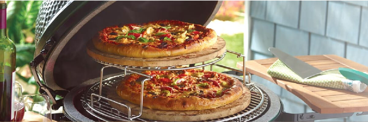 Two freshly baked pizzas with various toppings cooking on a multi-level rack inside a ceramic kamado-style grill on a patio table.