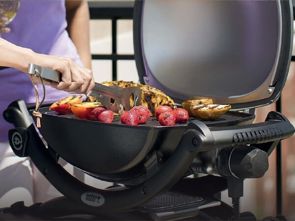Person grilling a mix of strawberries and vegetables on a compact electric grill with the lid open on a patio setting.