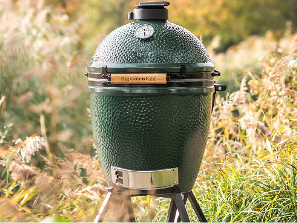 Large green ceramic grill with thermometer on top standing outdoors amid tall grass and soft-focus natural background.