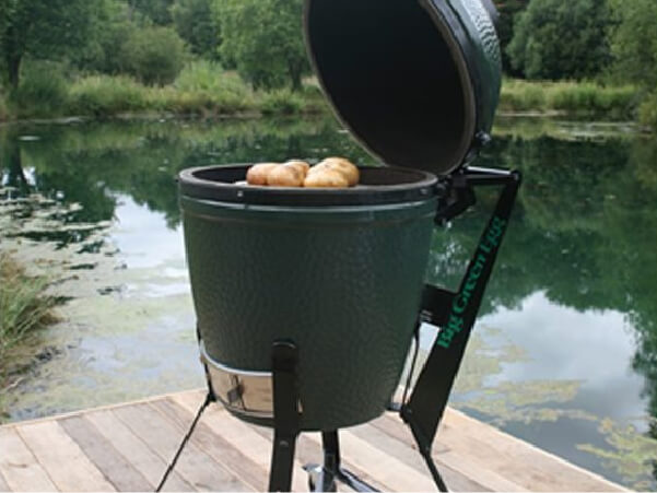 Green ceramic charcoal grill with the lid open cooking potatoes, positioned on a wooden dock by a calm lake surrounded by trees.