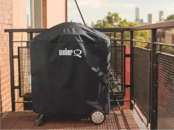 Black protective cover with white branding covering a round grill on a balcony with metal railings and a cityscape in the background.