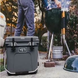 Gray portable cooler with a black YETI logo, sitting outdoors near a charcoal grill with a person preparing to cook in a backyard setting.