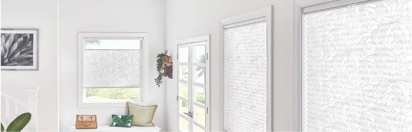 Bright interior corner with white textured roller shades covering multiple windows, a bench with cushions, a handbag, and a framed black-and-white photo on the wall.