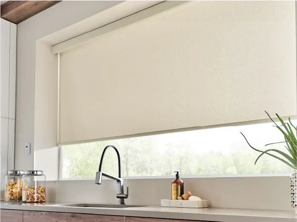 Modern kitchen sink area with a sleek black faucet, white countertop, wood cabinetry, clear storage jars, cream roller shade, and a small potted plant.
