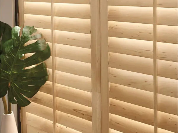 Close-up of light wood plantation shutters with natural wood grain texture, next to a white vase holding a large green leafy plant.