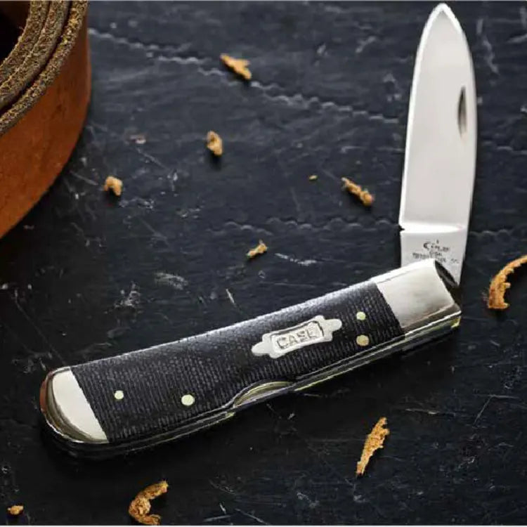 Open folding pocket knife with a black textured handle and polished stainless steel blade resting on a dark surface with small wood shavings nearby.