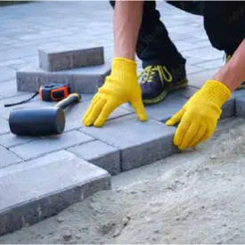 Person wearing bright yellow gloves placing rectangular gray paving stones on sand during an outdoor home improvement task.